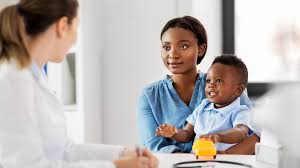A mother holds her smiling baby on her lap during a doctor's visit. The doctor listens attentively and translates with BAU a[[ by Techmed Connect. The atmosphere is caring and supportive.
